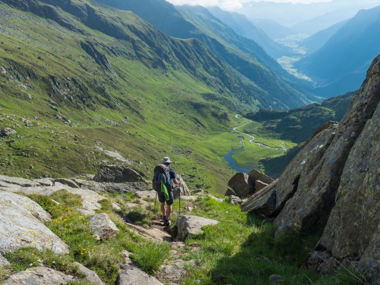 Ensam man vandrare med tung ryggsäck på Stubai vandringsled, Stubai Hohenweg på grön sommar alpina bergsdal med slingrande flod källström. Tyrolen, Österrikiska Alperna