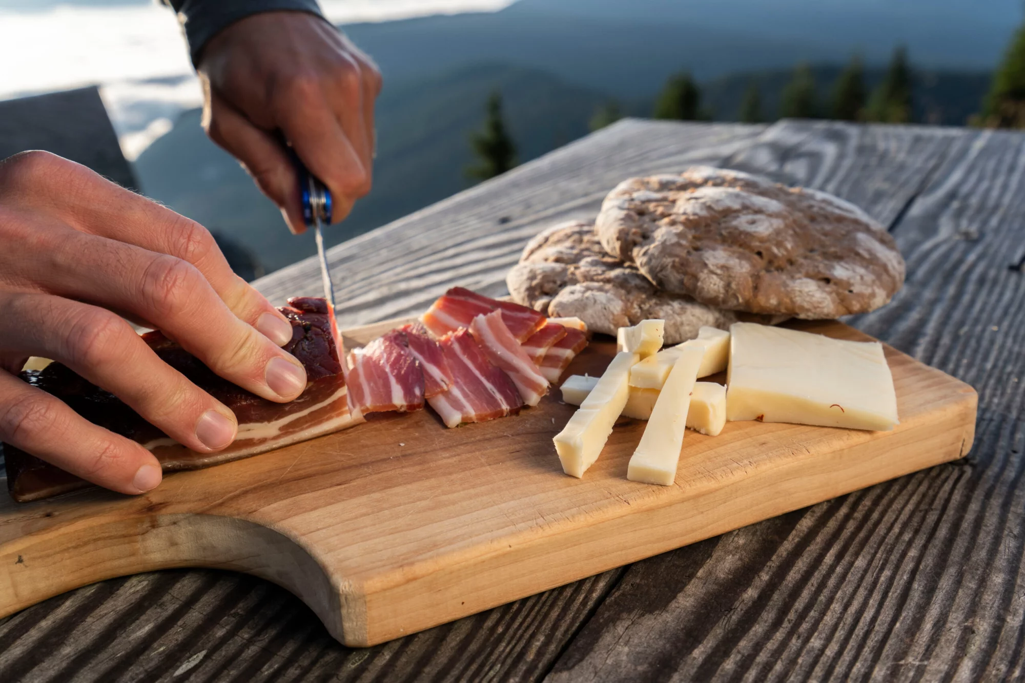 Un hombre corta delicioso tocino ahumado y queso del Tirol del Sur con pan plano sobre una mesa rústica.